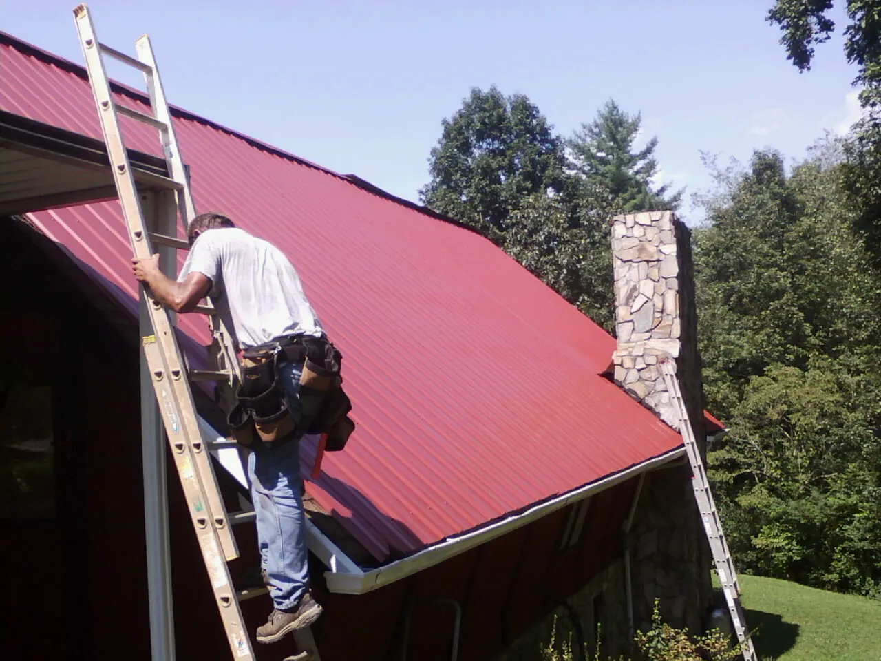 Roofer on a ladder beside a red metal roof and stone chimney — Soffit & Fascia Repair by Roofing Brothers in Saint Hilaire
